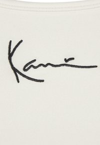 Biely textil s čiernym vyšitým podpisovým dizajnom. Materiál je hladký s ryhovanou textúrou a výrazným šitím.