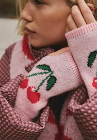 Rožinis megztas megztinis su raudonais ir žaliais vyšnių raštais, uždėtas ant languotos bluzelės, su tekstūruota medžiaga ir raštuotais kraštais.