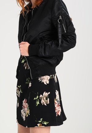 Vrouw die een zwarte bomberjas draagt over een zwarte bloemenrok met roze en witte bloemen, staand tegen een effen achtergrond.