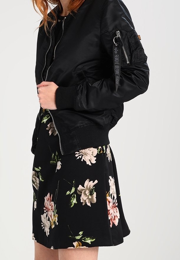 Femme portant une veste bomber noire sur une jupe noire à fleurs roses et blanches, debout devant un fond uni.