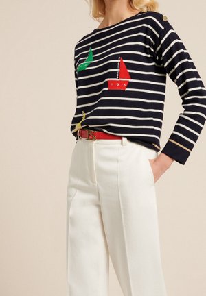 Pull rayé marin avec des appliqués de voiliers colorés, boutons dorés sur l'épaule, associé à un pantalon blanc taille haute et une ceinture rouge.