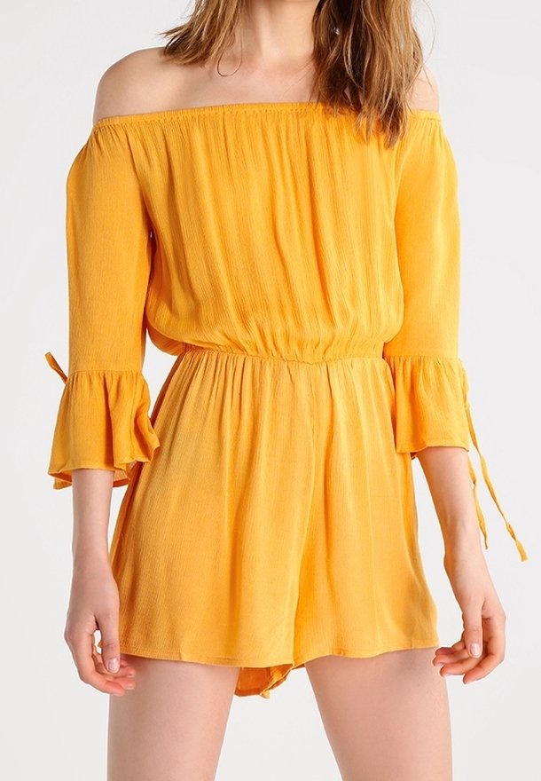 Femme portant une combinaison jaune épaules dénudées avec taille élastique et manches cloches trois-quarts nouées avec de petits nœuds.