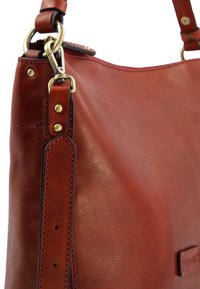 Sac à épaule en cuir rouge avec une texture lisse, une forme arrondie, une sangle renforcée et des accents en laiton. Détails cousus sur les bords.