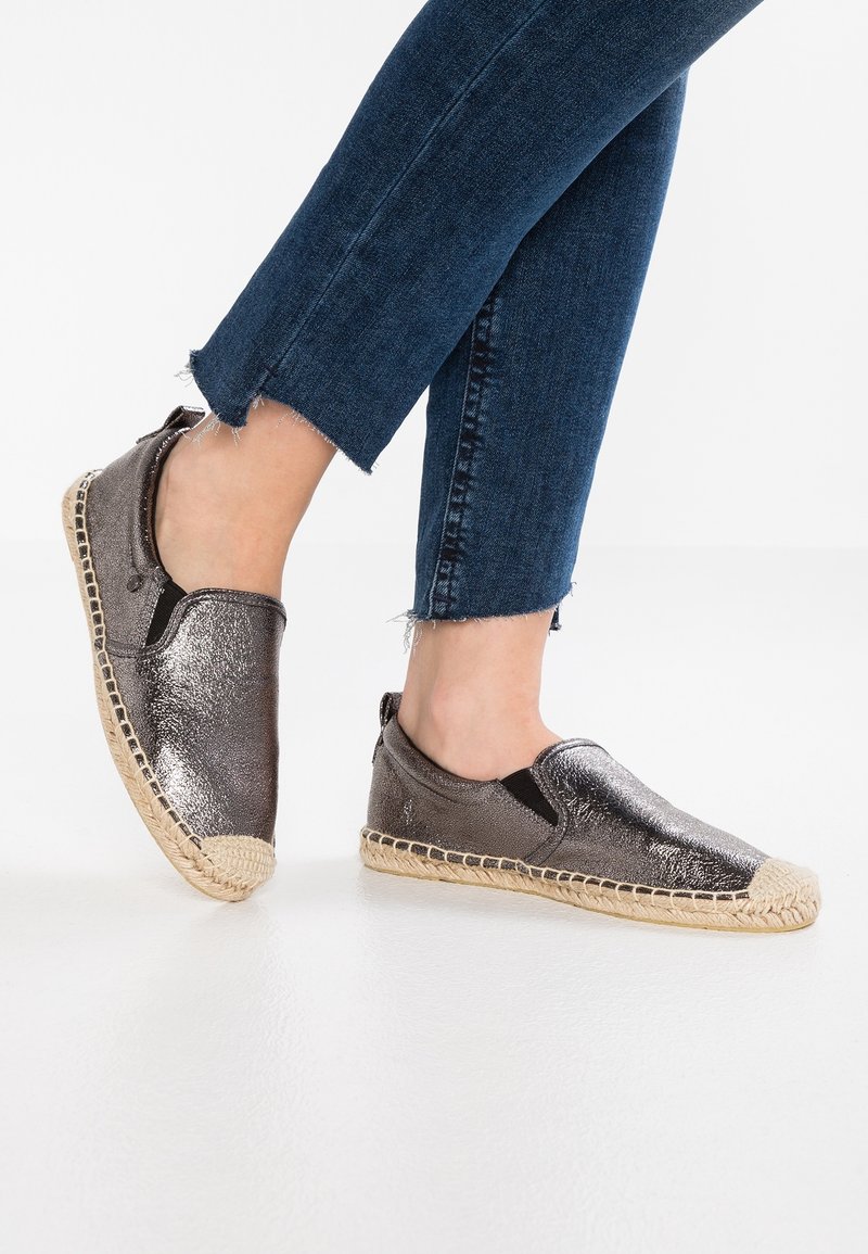 Pieds chaussés d'espadrilles métalliques à enfiler avec des semelles en jute tressé, portées avec un jean denim bleu foncé raccourci sur un sol blanc.