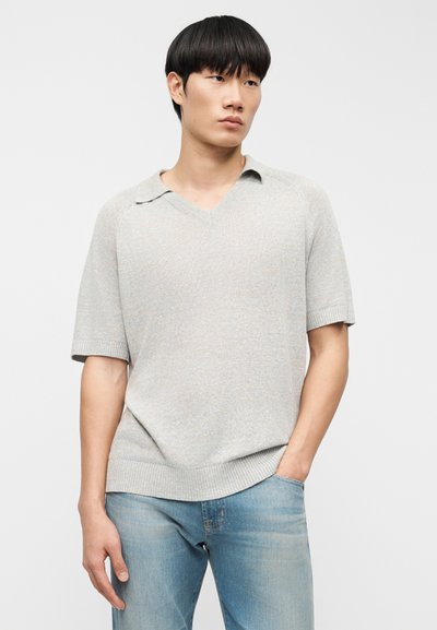 Νέος άνδρας με ίσια μαύρα μαλλιά, που φοράει ένα ανοιχτό γκρι πλεκτό πόλο με κοντά μανίκια και ανοιχτό μπλε τζιν, στέκεται με το ένα χέρι στην τσέπη.