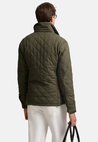 Giacca trapuntata verde oliva con collo alto, chiusura laterale a scatto e polsini elasticizzati. Indossata con pantaloni chiari e con una borsa nera in mano.