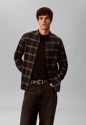 Giovane uomo che indossa una camicia a quadri nera con bottoni sopra una t-shirt nera, con pantaloni neri e cintura nera, in piedi con le mani nelle tasche.