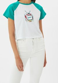 Bílý zkrácený tričko s tyrkysovými raglánovými rukávy. Na přední straně se nachází barevná retro grafika televizoru s nápisem "peace" uprostřed. Bavlněný materiál.