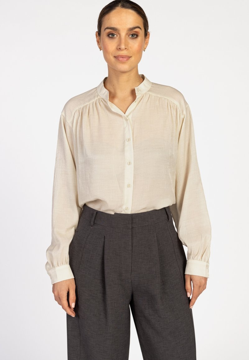 Light beige button-up shirt with a mandarin collar, long sleeves, and subtle texture, paired with high-waisted dark gray pleated trousers.