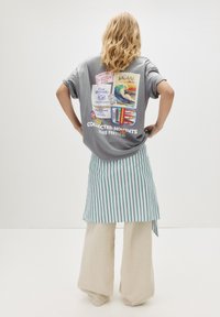 Person med langt blondt hår står med hænderne på hoften iført en grå graphic T-shirt, stribet omslagsskjorte og beige vidde bukser på hvid baggrund.