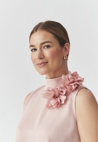 Bluza blijedo ružičaste boje s visokim ovratnikom, ukrašena slojevitim cvjetovima od tkanine na ramenu, s glatkom teksturom i bez rukava. Naglašena zlatna naušnica.