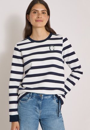 Kvinde iført en hvid og marineblå langærmet trøje med horisontale striber og blå jeans, stående med den ene hånd i lommen.