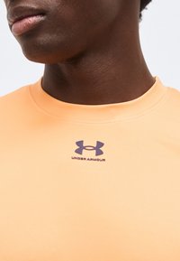 Κοντινό πλάνο ενός ατόμου που φοράει ροδακινί μπλουζάκι Under Armour με λογότυπο στο κέντρο κάτω από τον λαιμό σε απλό ανοιχτό φόντο.