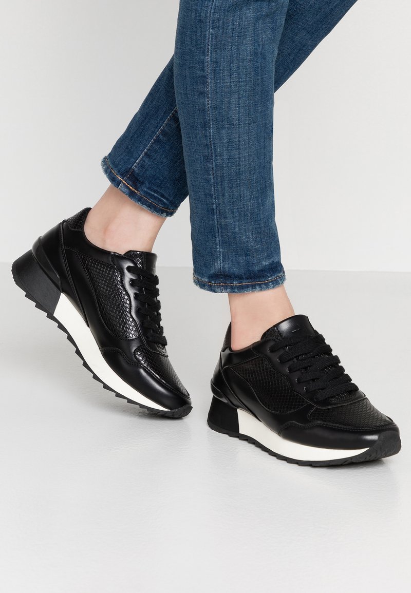 Black sneakers made of smooth and textured materials, featuring a white sole, black laces, and a rounded toe. Worn with blue jeans.