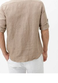 Hombre vistiendo una camisa de lino beige de manga larga con las mangas arremangadas y pantalones blancos, visto desde atrás sobre un fondo blanco.