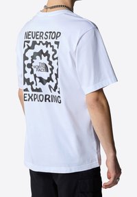 Biała bawełniana koszulka z czarną wirującą grafiką i napisem "NIGDY NIE PRZESTAWAJ ODKRYWAĆ" oraz logo "THE NORTH FACE" na plecach.