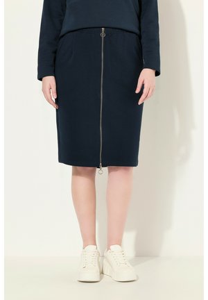 Jupe bleu marine longueur genou en tissu doux avec une fermeture éclair devant et une taille élastique, portée avec des baskets compensées blanches.