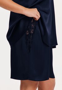 Robe de nuit en satin bleu marine avec détail en dentelle sur le côté, présentant une texture lisse et une coupe décontractée.