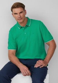 Groene polo met witte rand op de kraag en mouwen, korte mouwen, en een subtiel logo op de borst. Draag met donkere broek.