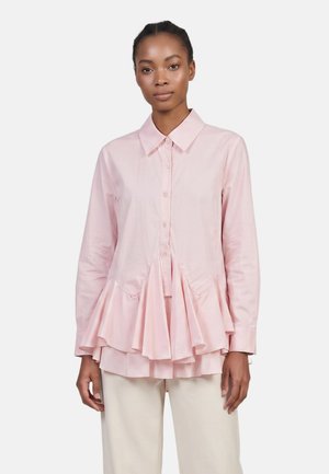 Femme portant une blouse rose clair boutonnée avec un ourlet volanté et des manches longues, associée à un pantalon beige clair, debout devant un fond uni.