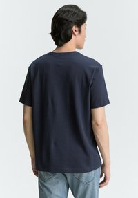 Dunkelblaues T-Shirt mit kurzen Ärmeln, geripptem Stoff, rundem Halsausschnitt und geradem Schnitt. Trägt eine helle blaue Jeans. Rückansicht.