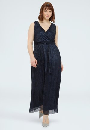 Mujer de cabello rojo con un vestido maxi azul marino sin mangas y textura, y tacones puntiagudos grises, de pie contra un fondo claro y liso.