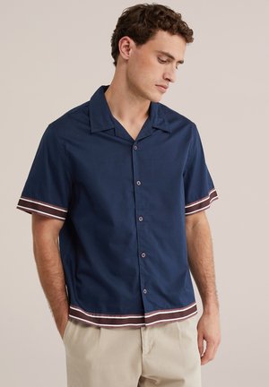 Homme portant une chemise bleu marine à manches courtes avec boutons, avec des détails à rayures marron et blanc sur les manches et l'ourlet, associée à un pantalon beige.