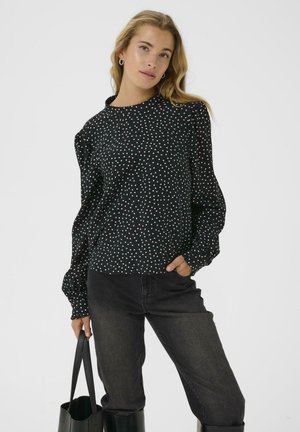 Schwarze Bluse mit langen Ärmeln und weißen Punkten, Rundhalsausschnitt und gerafften Ärmeln. Kombiniert mit hochtaillierten schwarzen Jeans und einer Tragetasche.