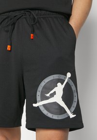 Shorts de sport noirs à texture en mesh, arborant un grand logo blanc Air Jordan et un graphisme circulaire. Cordon de serrage avec embouts orange.