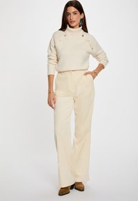 Cremfarbener Rollkragenpullover mit Knopfdetails, kombiniert mit hellbeigen, weiten Cordhosen und braunen, knöchelhohen Stiefeln mit Absatz.