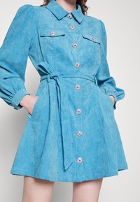 Femme portant une robe en velours côtelé bleu avec des boutons fleurs en cristal, des manches longues bouffantes, des poches avant et une ceinture nouée à la taille.