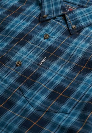Chemise à carreaux bleue avec des lignes bleu foncé, bleu clair et orange, dotée d'un col boutonné et d'une poche poitrine. Étiquette de marque "Superdry" visible.