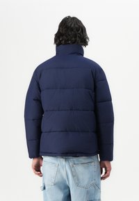 Marineblauwe gewatteerde jas met een hoge kraag en een gestikte textuur, voorzien van horizontale secties en elastische manchetten. Draag het met lichtblauwe spijkerbroeken.