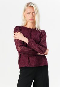 Burgundy blouse with textured leaf pattern, puffed sheer sleeves, and elastic cuffs, paired with black pants. Smooth, lightweight fabric.