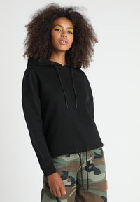 Felpa nera con cappuccio e coulisse, caratterizzata da una vestibilità ampia e un tessuto testurizzato. Abbinata a pantaloni con motivo mimetico.
