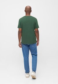 Groene T-shirt met korte mouwen en een ronde halslijn, gecombineerd met blauwe spijkerbroeken en witte sneakers, gezien vanuit de achterkant.
