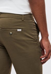 Olijfgroene broek van katoen met een plat achterzakje en knoop. Heeft een wit label met tekst op het zakje. Soepele textuur.