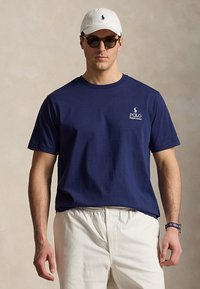 Άνδρας που φοράει μπλε μπλουζάκι Polo Ralph Lauren, λευκό παντελόνι, λευκό καπέλο, σκουρόχρωμα γυαλιά ηλίου και ρολόι χειρός Polo με μονόχρωμο φόντο.