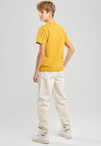 Camiseta amarilla de manga corta combinada con pantalones blancos de corte amplio y zapatillas blancas, vista desde la parte de atrás contra un fondo liso.