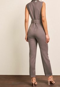 Mono gris sin mangas con cinturilla ajustada, piernas estrechas y diseño minimalista. Tela suave con un brillo sutil. Vista trasera.