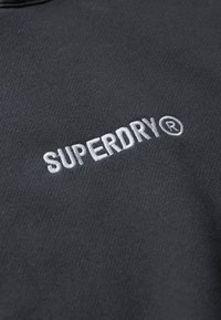 Sudadera negra con un logo "SUPERDRY" bordado en blanco. El material parece texturizado, con un cuello redondo y un diseño informal.