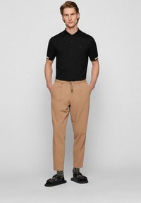 Svart polo skjorta med korta ärmar, beige avsmalnande byxor och svarta sandaler i kombination med mönstrade strumpor. Enkel design med minimala detaljer.