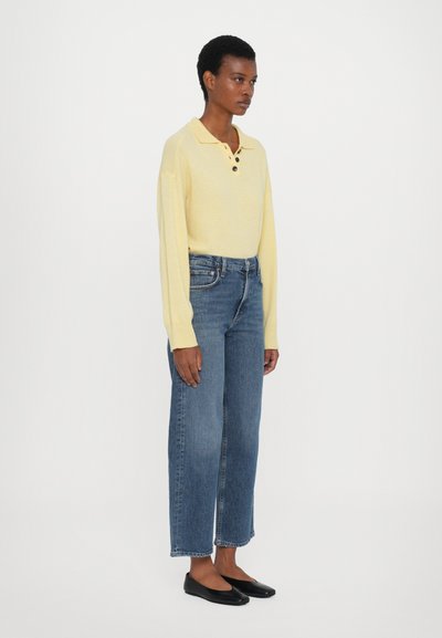 Geltona ilgomis rankovėmis polo marškinėliai su dviem mygtukais, derinami su mėlynomis tiesiomis džinsinėmis kelnėmis ir juodais baleto bateliais. Paprastas, švarus dizainas su laisvu prigludimu.