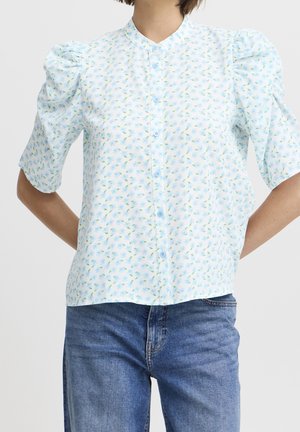 Woman wearing a white short-sleeve blouse with blue floral pattern and puffed shoulders, paired with blue denim jeans.