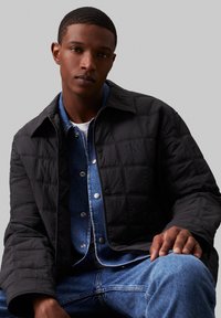 Jachetă neagră cu umplutură, cu guler, purtată peste o cămașă din denim albastru. Model notabil de cusături și logo subtil pe jachetă.