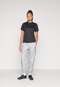 Juoda trumpomis rankovėmis Nike marškinėliai, derinami su šviesiai pilkomis sportinėmis kelnėmis su elastingais rankogaliais ir kišenėmis, užbaigiant juodais sportiniais batais.