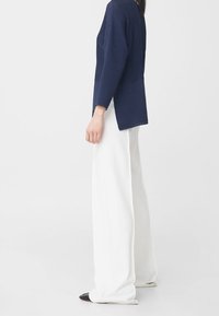 Pull en jersey bleu marine à côtes avec des manches longues, présentant un ourlet arrondi. Associé à un pantalon large blanc et des mocassins noirs à bout pointu.