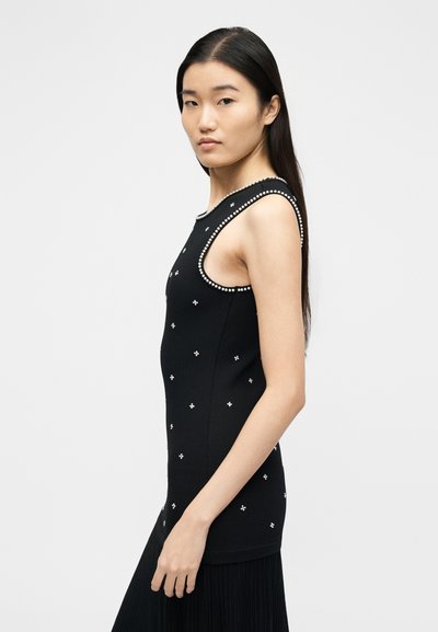 Νεαρή γυναίκα με μακριά σκούρα μαλλιά, που φοράει στενό μαύρο αμάνικο τοπ με μικρές λευκές διακοσμήσεις και μαύρη φούστα, στραμμένη προς τα πλαϊνά.