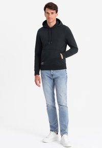 Felpa nera con tasca a marsupio, cappuccio con cordino e polsini a costine, abbinata a jeans di denim blu chiaro e scarpe da ginnastica bianche.