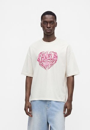 Νεαρός άνδρας φοράει λευκό oversized μπλουζάκι με ροζ καρδιά και ανοιχτό μπλε τζιν, στέκεται μπροστά σε απλό λευκό φόντο.